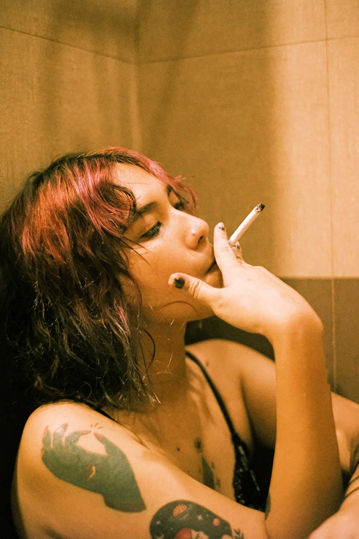 A woman with tattoos and unique hairstyle relaxes indoors, smoking a cigarette.